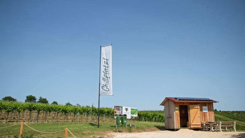 Rebenhof, Chillplatzl, © Rebenhof Ein kleiner Holzschuppen neben einem Weinberg mit einem Schild und einer Fahne mit der Aufschrift 'Chillplatz'.