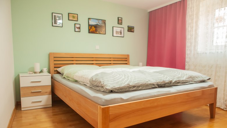 Schlafzimmer, © Fam. Heinrich Ein Schlafzimmer mit einem Holzbett, grüner Wand, Bildern, Nachttisch und rosa Vorhängen.