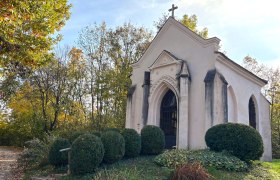 Kapelle, © "Natur im Garten" Eine kleine Kapelle mit gotischen Fenstern und einem Kreuz auf dem Dach, umgeben von Bäumen und Sträuchern im Herbst.