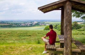 Panoramablick, © Fotostudio Semrad Ein Mann sitzt unter einem Holzunterstand und blickt auf eine weite Landschaft mit Feldern und einem Dorf in der Ferne.