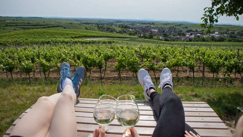 Rebenhof, Chillplatzl, © Rebenhof Zwei Personen entspannen auf einer Bank in einem Weinberg mit Blick auf die Landschaft, während sie Weingläser anstoßen.