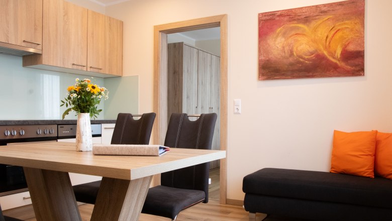 Rebenhof, Ferienwohnung, © Rebenhof Moderne Küche mit Holzmöbeln, Esstisch, Stühlen und einem Sofa mit orangefarbenen Kissen.