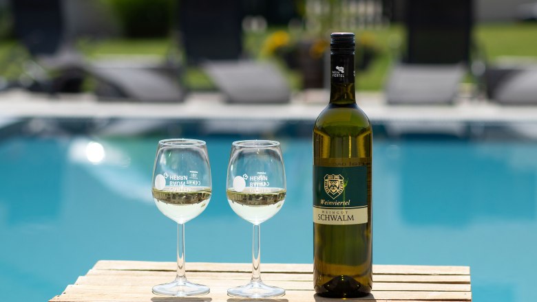 Rebenhof, Ferienwohnung & Weingut, © Rebenhof Eine Flasche Wein und zwei Weingläser auf einem Holztisch vor einem Pool.