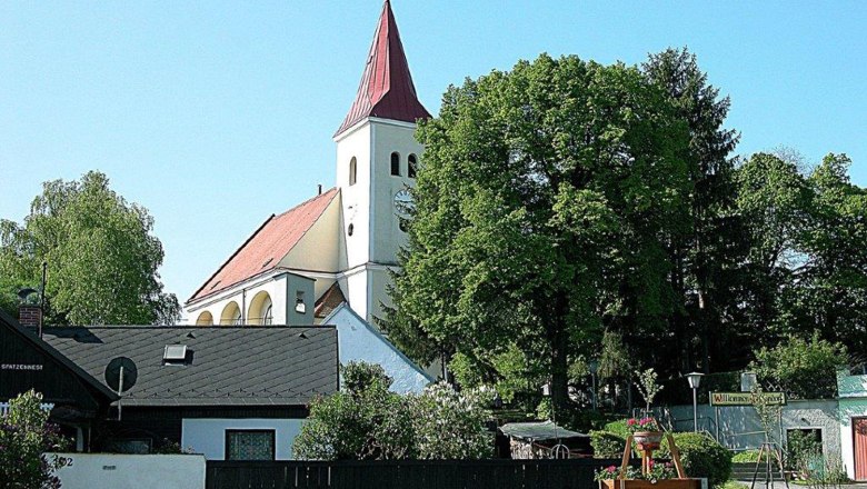 Kirche, © Gemeinde Hauskirchen Eine Kirche mit rotem Dach und Turm, umgeben von Bäumen und Häusern.