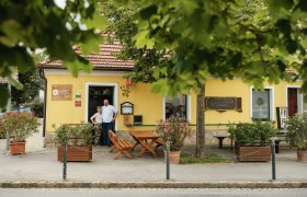 Gasthaus Herbst, © Weinviertel Tourismus / Michael Reidinger Ein gelbes Gasthaus mit rotem Dach, umgeben von Bäumen. Zwei Personen stehen vor dem Eingang.