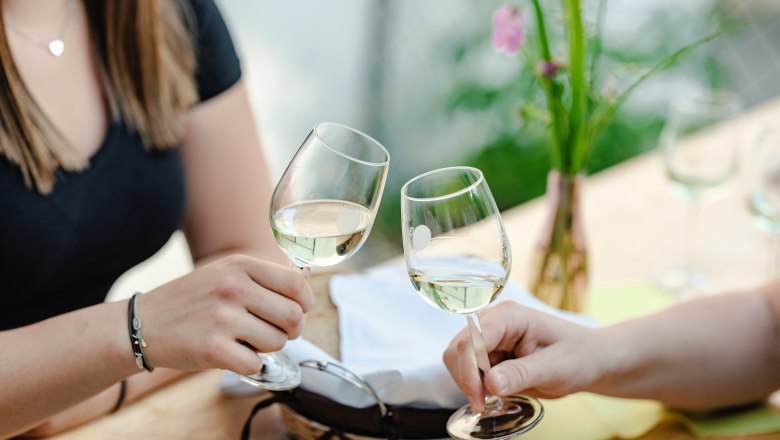 Prost, © Michael Reidinger Zwei Personen stoßen mit Weißweingläsern an einem Tisch an.