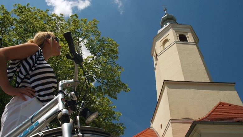 Wallfahrtskirche Maria Moos, © Stadtgemeinde Zistersdorf Eine Frau mit Fahrrad steht vor einem Kirchturm und blickt nach oben.