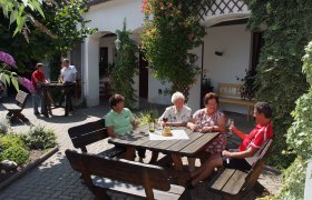Arkadenhof-Heuriger, © Seymannfilm Menschen sitzen an einem Tisch im Freien und genießen Getränke in einem begrünten Innenhof.