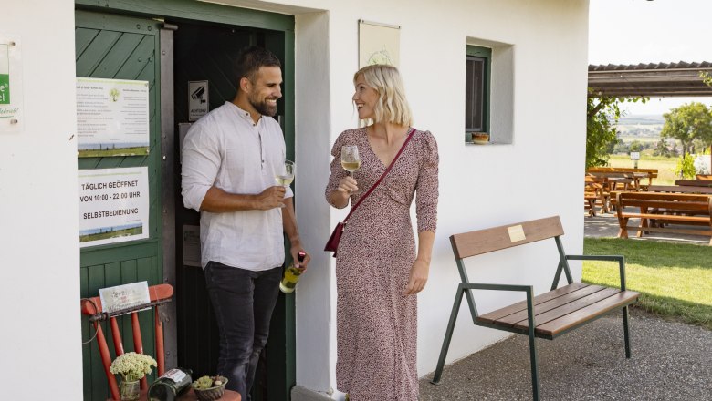 Selbstbedienungskeller beim Baumkreis, © Weinviertel Tourismus GmbH / Schwarz-König Ein Mann und eine Frau stehen vor einem kleinen weißen Gebäude und trinken Wein.
