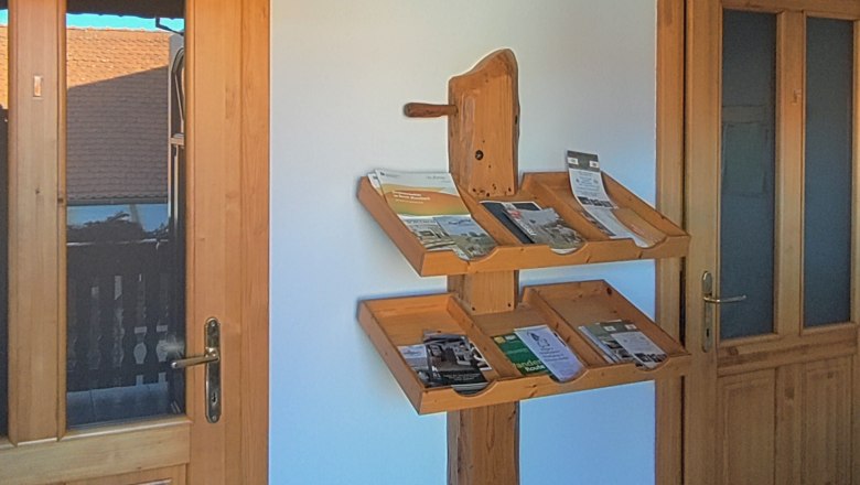 Große Auswahl an Prospekten, © Arkadenhof Schneider Ein Holzregal mit Prospekten an einer weißen Wand zwischen zwei Holztüren.