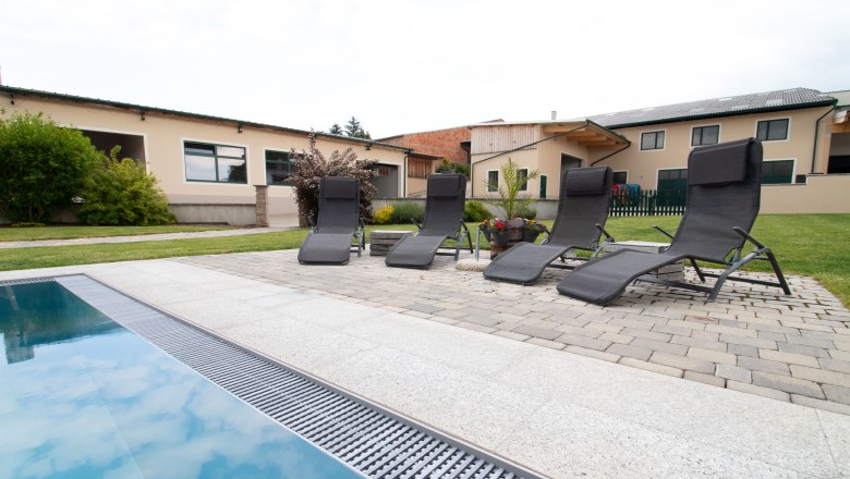 Rebenhof, Ferienwohnung, © Rebenhof Vier Liegestühle stehen neben einem Pool auf einer Terrasse mit Pflastersteinen. Im Hintergrund sind Gebäude und ein Garten zu sehen.