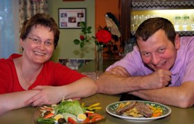 Familie Herbst, © Seymannfilm Ein Mann und eine Frau lächeln in die Kamera, vor ihnen stehen Teller mit Essen. Im Hintergrund ist eine rote Rose und ein Regal mit Gläsern zu sehen.