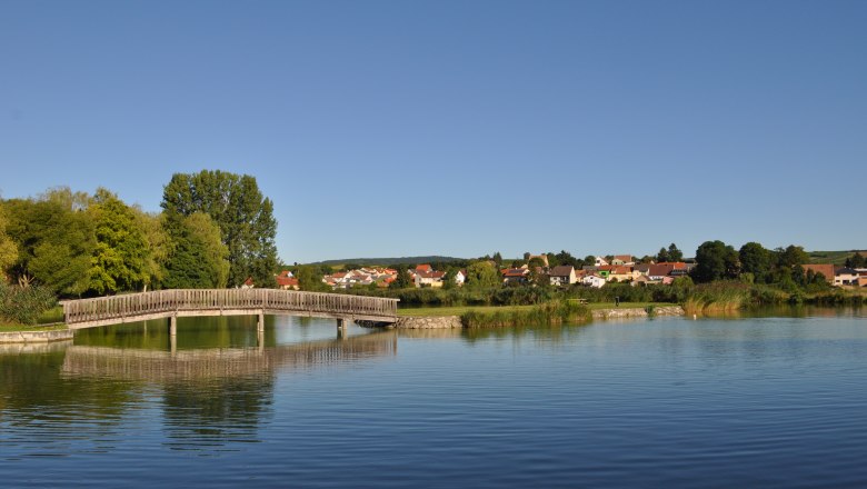 Badeteich Poysdorf, © Weinviertel Tourismus Holzbrücke über einen ruhigen Teich mit Dorf im Hintergrund.