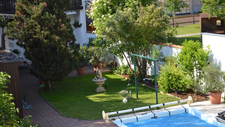 Garten, © Karoline Schmidt Ein gepflegter Garten mit Bäumen, Sträuchern, einer Statue und einem Pool. Im Hintergrund sind ein Haus und ein Zaun zu sehen.