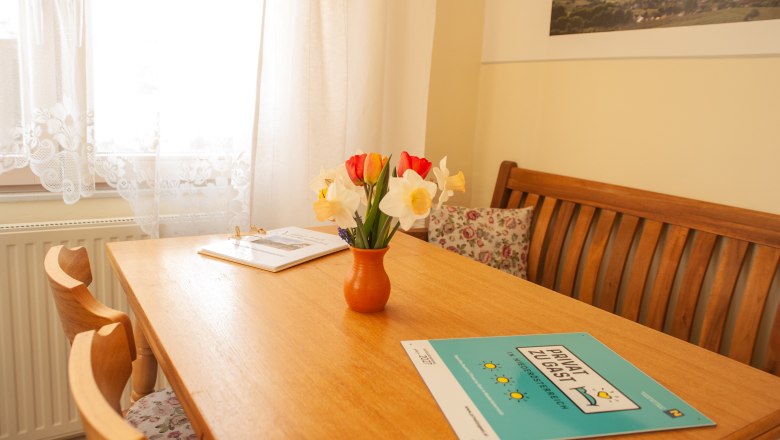 Essecke, © Fam. Heinrich Ein gemütlicher Essbereich mit Holztisch, Blumenstrauß in Vase und Broschüren. Helle Vorhänge und ein Bild an der Wand.