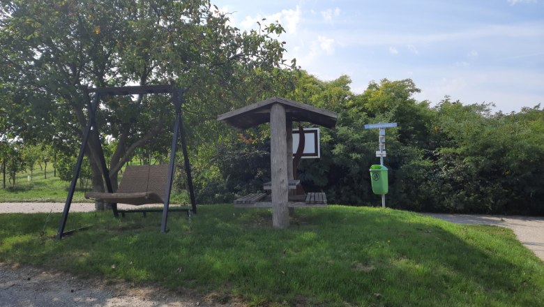 Weinriedenrastplatz Gaubitsch, © Gemeinde Gaubitsch Ein Rastplatz mit einer Schaukel und einem überdachten Sitzbereich neben einem Baum und einem Mülleimer auf einer Wiese.