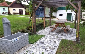 Trinkbrunnen Rastplatz Erdberg, © Vino Versum Poysdorf Ein Rastplatz mit Trinkbrunnen, Holztisch und Sitzbank unter einer Pergola, umgeben von grüner Wiese und kleinen Gebäuden.
