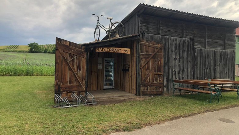 Eine rustikale Holzh&uuml;tte mit der Aufschrift 'Radlerrast' und einem Fahrrad auf dem Dach, umgeben von Feldern und einer Bank.