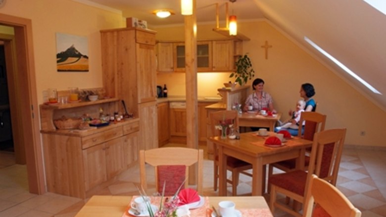 Ein gem&uuml;tlicher Fr&uuml;hst&uuml;cksraum mit Holzm&ouml;beln, gedeckten Tischen und zwei Personen, die sich unterhalten. Ein Kind sitzt auf dem Scho&szlig; einer Person.