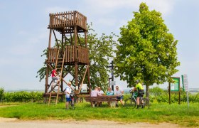 Menschen entspannen an einem Holzturm und Tisch im Freien, umgeben von B&auml;umen und Schildern.