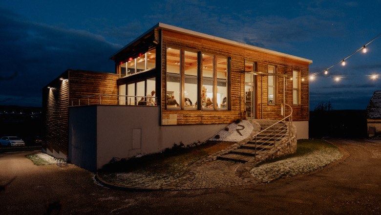 Außenansicht eines modernen Heurigenlokals bei Nacht mit beleuchteten Fenstern und Lichterkette.