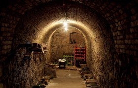 Ein schmaler, gew&ouml;lbter Weinkeller mit Ziegelw&auml;nden und gelagerten Flaschen.
