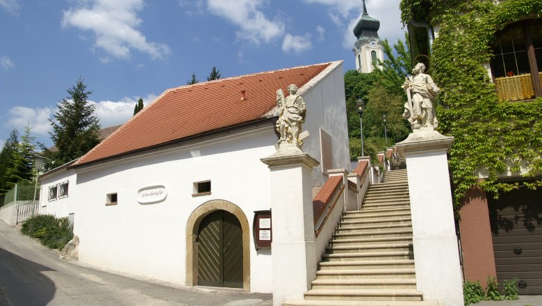 Weißes Gebäude mit rotem Dach und Treppe mit Statuen, Kirche im Hintergrund.