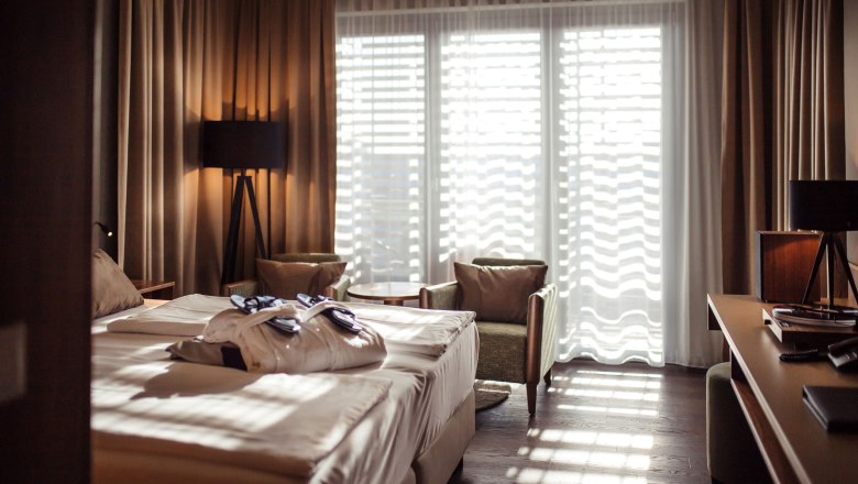 Hotelzimmer mit Doppelbett, Bademänteln, zwei Sesseln und einem Tisch vor einem großen Fenster mit Vorhängen.