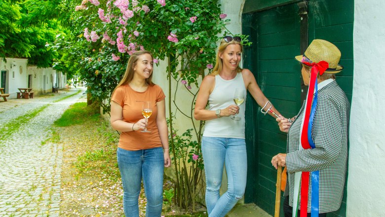 Drei Personen stehen vor einem gr&uuml;nen Tor, zwei halten Weingl&auml;ser, eine tr&auml;gt einen Strohhut mit bunten B&auml;ndern.