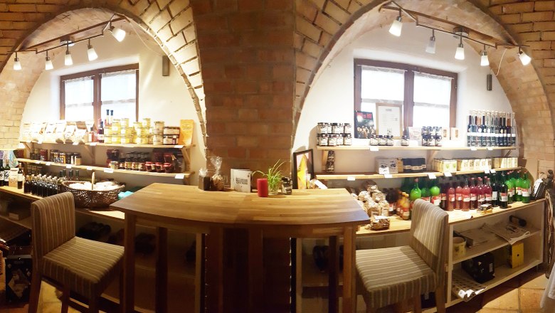 Innenansicht eines kleinen Ladens mit Regalen voller Lebensmittel und Getränken, Holztisch und Stühlen.
