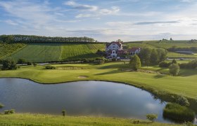 Landschaftsaufnahme eines Golfplatzes mit Teich und Clubhaus im Hintergrund.
