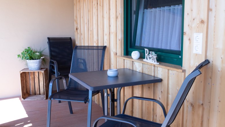 Ein Balkon mit Holzwänden, zwei schwarzen Stühlen, einem Tisch und einer Pflanze in einem Topf.