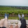 Zwei Personen entspannen auf einer Bank in einem Weinberg mit Blick auf die Landschaft, w&auml;hrend sie Weingl&auml;ser ansto&szlig;en.