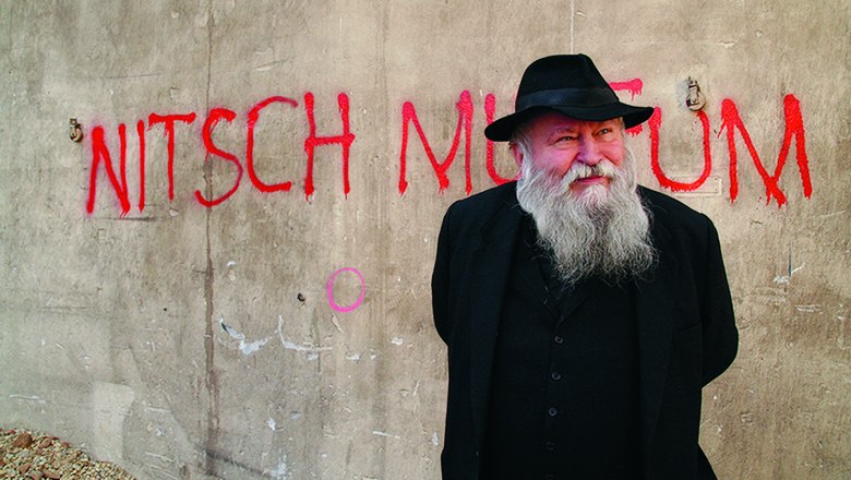 Ein Mann mit Bart und Hut steht vor einer Wand mit der Aufschrift 'Nitsch Museum'.