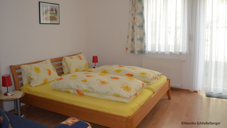 Ein gemütliches Doppelzimmer mit einem Holzbett, bunter Bettwäsche und zwei roten Nachttischlampen. Ein Fenster mit Vorhängen ist im Hintergrund.