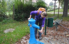 Eine Person f&uuml;llt eine Wasserflasche an einem blauen Trinkbrunnen im Freien auf.