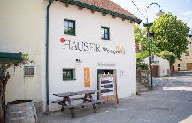 Au&szlig;enansicht eines Chalets mit der Aufschrift 'Hauser Weingenuss'. Vor dem Geb&auml;ude steht ein Holztisch und eine Tafel mit Informationen.
