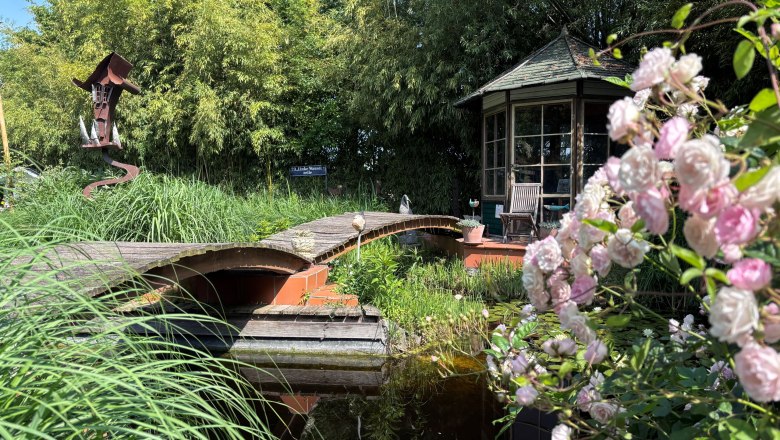 Ein idyllischer Garten mit Teich, Brücke, Pavillon und blühenden Rosen im Vordergrund.