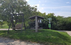 Ein Rastplatz mit einer Schaukel und einem &uuml;berdachten Sitzbereich neben einem Baum und einem M&uuml;lleimer auf einer Wiese.