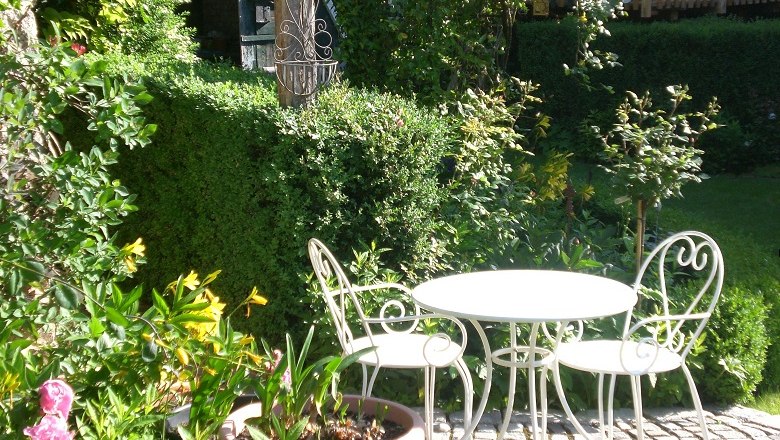 Ein idyllischer Garten mit einem weißen Metalltisch und zwei Stühlen auf einem gepflasterten Bereich, umgeben von grünen Pflanzen und Sträuchern.
