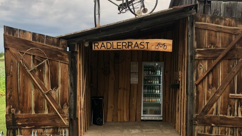 Holzh&uuml;tte mit Schild 'Radlerrast', Fahrrad auf dem Dach, Getr&auml;nke im Inneren.