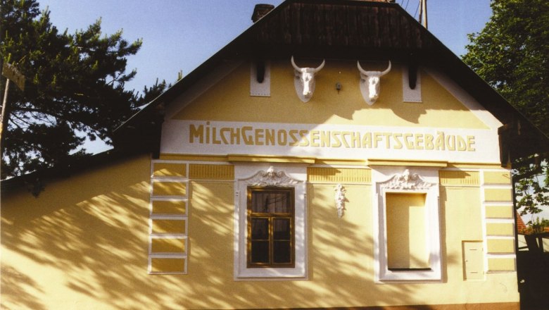 Gelbes Gebäude mit der Aufschrift 'Milchgenossenschaftsgebäude' und zwei Stierköpfen an der Fassade.