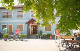 Ein blaues Gasthaus mit Sonnenschirmen und Tischen im Freien, umgeben von grünen Bäumen.
