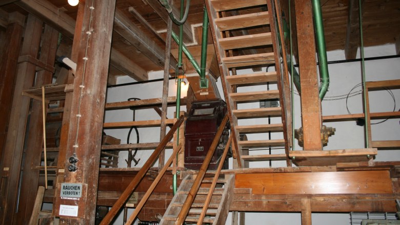 Innenansicht einer alten Mühle mit Holztreppen und Maschinen.