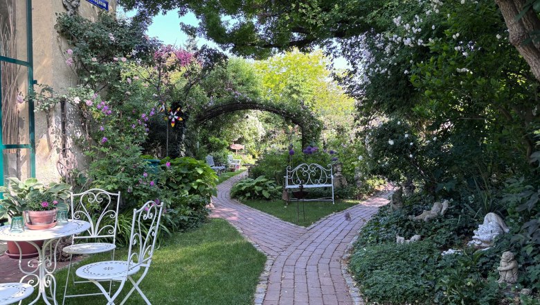 Ein idyllischer Garten mit weißen Gartenmöbeln, einem gepflasterten Weg und üppiger Vegetation.