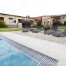Vier Liegest&uuml;hle stehen neben einem Pool auf einer Terrasse mit Pflastersteinen. Im Hintergrund sind Geb&auml;ude und ein Garten zu sehen.