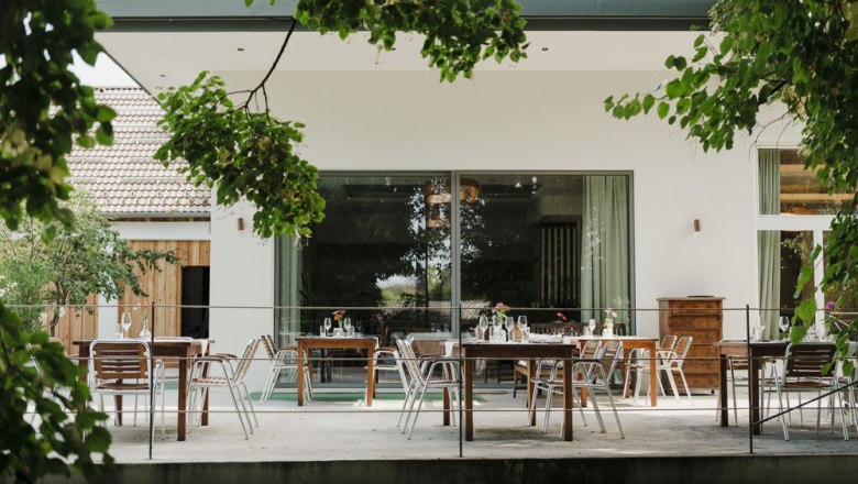 Terrasse mit Tischen und St&uuml;hlen, umgeben von gr&uuml;nen B&auml;umen.