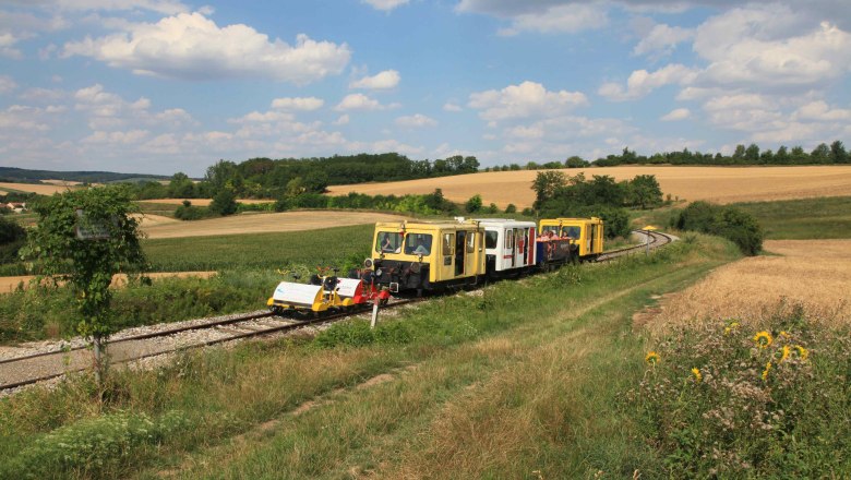 Draisinen auf einer l&auml;ndlichen Bahnstrecke in einer h&uuml;geligen Landschaft mit Feldern und B&auml;umen.