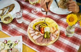 Ein gedeckter Tisch mit einer Platte voller Aufschnitt, Käse und Gemüse, dazu Weißwein und Brot auf einer rot-weiß karierten Tischdecke.