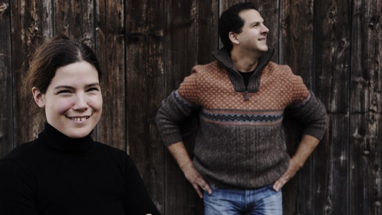 Zwei Personen vor einer Holzwand, eine Frau im Vordergrund lächelt, ein Mann im Hintergrund schaut zur Seite.